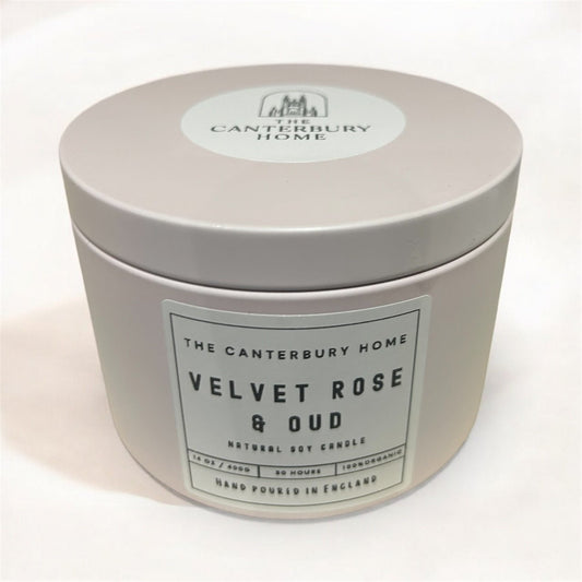 A lit three-wick candle in a pale pink round tin with its lid leaning beside it on a marble surface. The label reads 'Velvet Rose & Oud'.